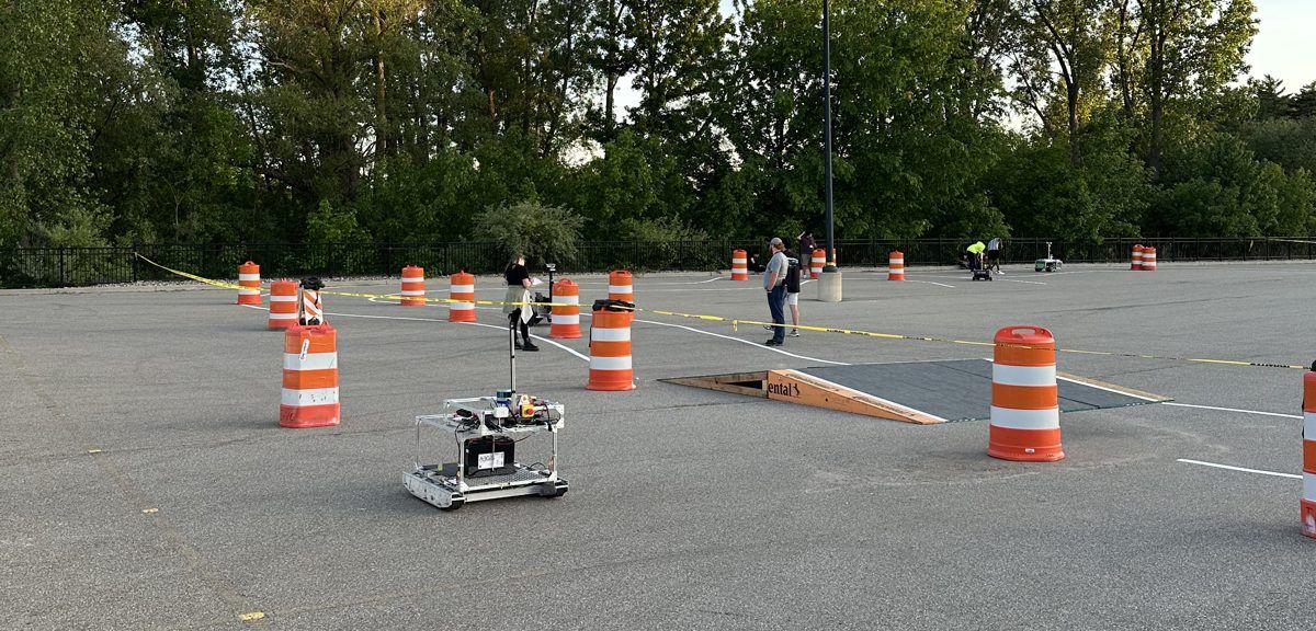 The IGVC 2026 course: an asphalt lot with orange construction barrels, painted lane lines and a plywood ramp, with a competing rover in the foreground.